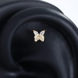 May include: Two gold-toned butterfly stud earrings featuring clear, marquise-cut stones arranged to form the wings. Each earring is secured in a black ear-shaped display stand, showcasing the detailed, symmetrical design of the butterfly shape.
