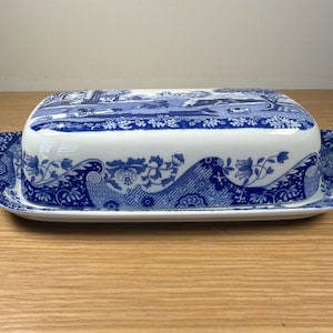May include: A rectangular ceramic butter dish featuring a white base with a blue transferware pattern. The lid and tray are decorated with intricate floral motifs and a detailed landscape scene depicting buildings and trees in a classic style.