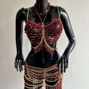 May include: A body jewelry set featuring a halter-style top and a waist chain made of deep red faceted beads and gold-toned metal chains. The top consists of multiple beaded strands connected by a central gold chain, while the waist piece features layered chains and beaded fringe accents.