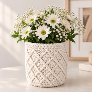 May include: A cylindrical off-white crochet basket featuring a raised diamond lattice pattern. The basket is filled with a bouquet of white daisies with yellow centers and small clusters of white baby's breath flowers with green stems.