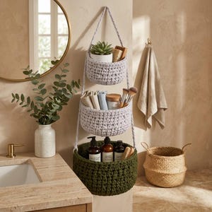 May include: A three-tiered hanging storage organiser featuring two light grey crocheted baskets and one dark olive green crocheted basket. The baskets are connected by braided cords and contain various bathroom items including a succulent and bottles.