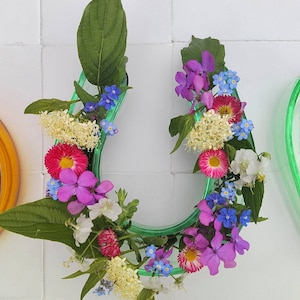 May include: A green glass vase shaped like a horseshoe filled with a variety of flowers, including pink, purple, white, and blue flowers.