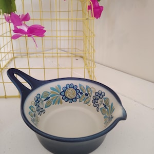 Puede incluir: Un cuenco de cerámica azul con asa y diseño floral en azul, verde azulado y dorado. El cuenco tiene un interior blanco y un pico vertedor. El fondo incluye una cesta de alambre amarilla y flores rosas.