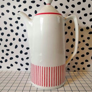 Puede incluir: Una cafetera de cerámica blanca con una franja roja alrededor de la parte superior y rayas verticales rojas cerca de la base. La cafetera tiene una tapa con un acento rojo y un asa curva.
