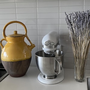 May include: A yellow and brown ceramic jug with a handle and lid, a white KitchenAid stand mixer with a stainless steel mixing bowl, and a clear glass vase filled with dried purple lavender stems arranged on a white kitchen counter.