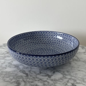 Puede incluir: Un cuenco de cerámica redondo con un patrón denso y repetitivo de pequeñas flores azules y enredaderas sobre un fondo blanco. El cuenco tiene un interior curvo y liso, con un borde azul oscuro sólido que define su contorno circular.