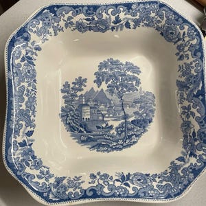 May include: A square ceramic bowl with a white base and blue transferware pattern. The rim features a detailed floral and scroll border. The center depicts a landscape scene with buildings, trees, mountains, and a small boat on the water.