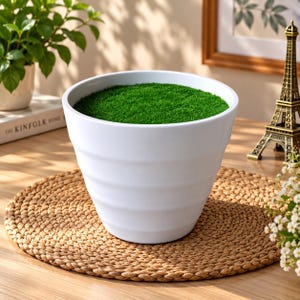 May include: A white ceramic planter with a tapered, ribbed exterior design. The interior of the pot is filled with a layer of vibrant green artificial grass, creating a contrast between the smooth white surface and the textured green top.