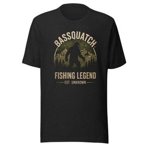 May include: A black short-sleeve t-shirt featuring a graphic print of a silhouette of a creature resembling Bigfoot standing in front of mountains and trees. The text reads BASSQUATCH FISHING LEGEND EST. UNKNOWN in a distressed beige font.