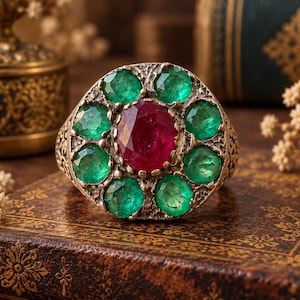 Peut inclure: Une bague ornée d'une grande pierre précieuse rouge foncé de taille ovale au centre, entourée de huit pierres précieuses vert vif de taille ronde. L'anneau en métal présente une finition sombre texturée avec des détails complexes.