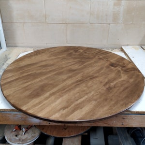 May include: A large, round wooden tabletop with a rich, brown stain. The wood grain is visible, creating a natural pattern across the surface. The tabletop is likely for a dining table or similar furniture piece.