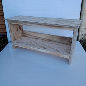 May include: A natural wood bench with a shelf. The bench is constructed from light-colored plywood, showcasing the wood grain. The design features a rectangular top and a lower shelf, supported by side panels.