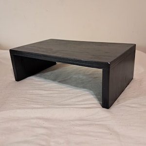 May include: A black wooden riser with a rectangular top and open sides. The riser is made of wood and has a smooth finish.