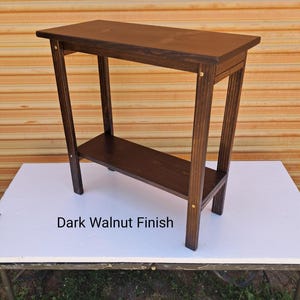 May include: A dark walnut finish console table with a lower shelf. The table has a rectangular top and a simple, modern design. The text "Dark Walnut Finish" is printed on the table.
