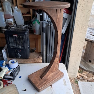 May include: A handcrafted wooden plant stand with a dark brown finish. The stand features a round top, a curved support, and a rectangular base. The stand is approximately 30 inches tall.