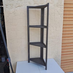 May include: A dark gray corner shelf with three triangular shelves. The wooden structure is designed to fit snugly into a corner, maximizing space. The shelves are evenly spaced, providing storage or display space.