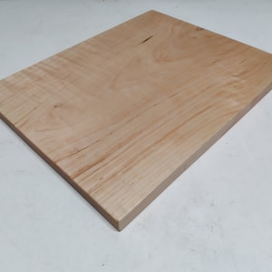 May include: A rectangular wooden cutting board with a light brown color and visible wood grain. The board has a smooth surface and a beveled edge. The wood appears to be maple or a similar hardwood.