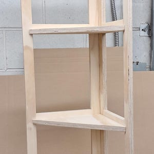 May include: A natural wood corner shelf unit with three shelves. The shelves are triangular and supported by a frame. The unit is made of light-colored plywood and is designed to fit into a corner.