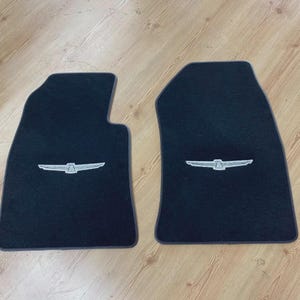 Può includere: Una coppia di tappetini neri per auto con un emblema ricamato color argento centrato che raffigura un'aquila stilizzata con le ali spiegate. I tappetini presentano una forma sagomata su misura progettata per adattarsi al pianale del veicolo.