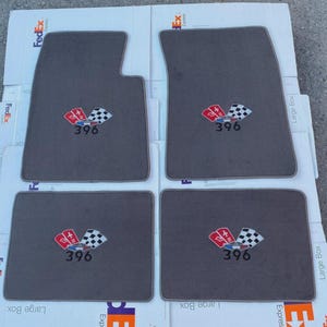 Könnte beinhalten: Ein Set aus vier dunkelgrauen Automatten mit einem gestickten Emblem, das eine rote Flagge, eine Zielflagge und die Zahl 396 in schwarzem Garn zeigt. Die Matten sind rechteckig, wobei eine Matte ein Design mit einer ausgeschnittenen Ecke aufweist.