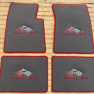 May include: A set of four dark gray automotive floor mats with red piping. Each mat features an embroidered logo with a red and white checkered flag, a leaping deer emblem, and the red text Impala SS centered on the carpeted surface.