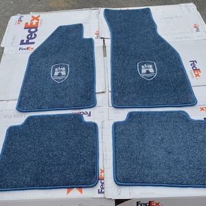 Puede incluir: Un juego de cuatro alfombrillas azules para coche con superficie texturizada y borde ribeteado en azul claro. Dos alfombrillas presentan un logotipo de escudo bordado en blanco que representa una torre de castillo sobre una ola.