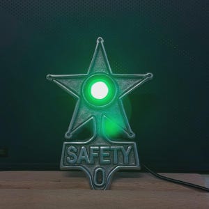 Peut inclure: Un emblème en métal argenté en forme d'étoile avec une lumière circulaire centrale brillant en vert. Sous l'étoile, une plaque rectangulaire affiche le texte en relief "SAFETY" au-dessus d'une petite découpe ovale. Un fin cordon noir dépasse.