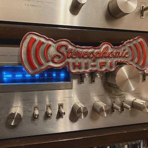 Puede incluir: Un letrero plateado y rojo con el texto Stereophonic HI-FI y líneas de señal curvas, colocado sobre un amplificador estéreo Pioneer plateado con una pantalla azul iluminada, perillas de control circulares e interruptores.