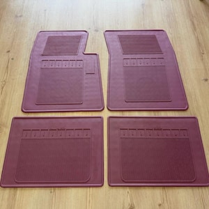 May include: A set of four maroon rubber car floor mats. The two front mats feature a distinct notched corner design, while the two rear mats are rectangular. Each mat has a textured surface with vertical ribbed patterns and a raised border edge.