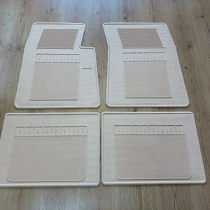 May include: A set of four beige automotive floor mats. The two front mats feature a distinct upper section with horizontal ridges and a larger lower section with vertical grooves. The two rear mats are rectangular with a matching vertical groove pattern.