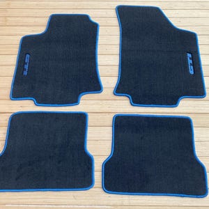 May include: A set of four black automotive floor mats with bright blue piping along the edges. Each mat features a textured carpet surface. The two front mats include a blue embroidered GTI logo positioned vertically near the outer edge.