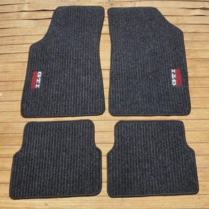 Puede incluir: Un juego de cuatro alfombrillas de coche de color gris oscuro con textura acanalada. Las dos alfombrillas delanteras más grandes tienen un parche rectangular con el texto blanco GTI y un cuadrado rojo. Las dos traseras tienen un diseño igual.