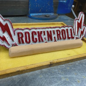 Könnte beinhalten: Ein gegossenes Metallschild mit der Aufschrift ROCK 'N' ROLL in roter Schrift und silbernen Blitzakzenten an den Enden. Das Schild steht hinter einem zylindrischen Holzdübel auf einem rechteckigen Holztablett mit einer gelben Unterlage.
