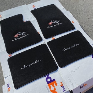 Könnte beinhalten: Ein Set aus vier schwarzen Automatten. Zwei Matten zeigen ein gesticktes Impala-Logo mit einem Zielflaggen-Emblem, während die anderen beiden Matten den Schriftzug Impala in weißer Schreibschrift zeigen. Die Matten haben eine flauschige Textur.