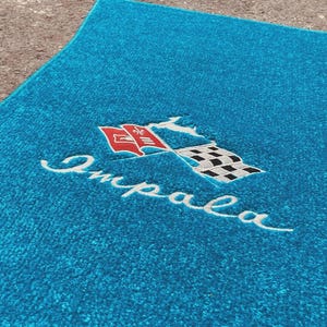 May include: A bright turquoise automotive floor mat featuring an embroidered logo with the word Impala in white script, a red flag with a white emblem, a black and white checkered flag, and a small white leaping impala silhouette graphic.