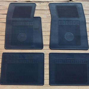 May include: A set of four black rubber car floor mats. The two front mats feature a distinct notched corner, a textured rectangular heel pad area, and a circular centre design. The two rear mats are rectangular with vertical ribbed patterns.