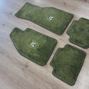 Peut inclure: Un ensemble de quatre tapis de sol en moquette vert olive pour véhicule. Chaque tapis présente un bord passepoilé vert foncé et un emblème central en relief en forme de bouclier. Le lot comprend deux tapis avant profilés et deux tapis arrière.