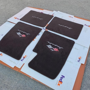 May include: A set of four dark brown automotive floor mats. Two mats feature an embroidered white script logo reading Impala above a checkered flag emblem. The other two mats are plain dark brown rectangles with the same white script logo.