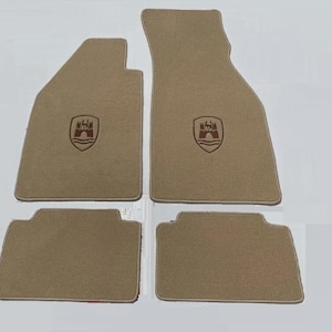 May include: A set of four beige automotive floor mats with a textured surface and bound edges. Each mat features a dark brown embroidered crest depicting a castle gate with three towers above wavy lines, centered on the upper portion of the carpet.