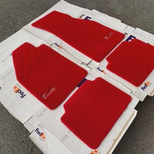 Peut inclure: Un ensemble de quatre tapis de sol automobiles rouge vif. Un tapis présente un motif matelassé et le mot Beetle brodé en écriture cursive. Un autre tapis présente le mot Beetle brodé en écriture cursive. Les tapis ont une texture pelucheuse.