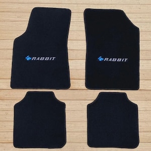 Puede incluir: Un juego de cuatro alfombrillas negras para coche. Las dos alfombrillas delanteras más grandes presentan un icono de conejo azul bordado seguido del texto blanco RABBIT. Las dos alfombrillas traseras más pequeñas son negras lisas.