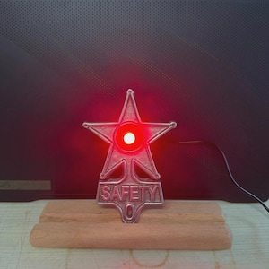 May include: A metallic star-shaped badge with a silver finish featuring the word SAFETY embossed at the bottom. A bright red light glows from the centre of the star. The badge is mounted upright on a rectangular light-coloured wooden base.