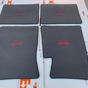 May include: A set of four dark grey automotive floor mats with black trim. Each mat features a circular emblem in the centre with the letters SS embroidered in red thread. The mats are arranged in a two-by-two grid on a flat surface.