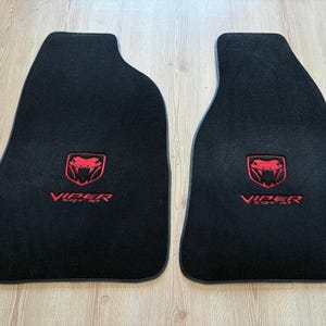 Könnte beinhalten: Ein Paar schwarze Automatten mit einem rot gestickten Viper SRT-10 Logo und Schlangenemblem. Die Matten haben eine konturierte Form für den Fahrzeuginnenraum, wobei der Schriftzug Viper SRT-10 deutlich in roter Stickerei zu sehen ist.