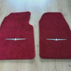 Puede incluir: Un par de alfombrillas de coche de color rojo intenso con una superficie texturizada y alfombrada. Cada alfombrilla presenta un emblema plateado centrado que representa un ave estilizada con las alas extendidas sobre el tejido rojo.