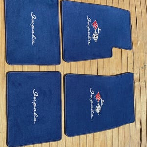 Op de afbeelding: Een set van vier donkerblauwe automatten. Twee matten zijn voorzien van het woord Impala in wit schrift, terwijl de andere twee het woord Impala in wit schrift bevatten naast een rood-wit geblokt vlag-embleem met een klein hert-silhouet.