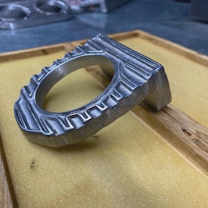 May include: A metallic, silver-coloured circular component featuring a textured, ridged outer edge and a flat, rectangular base section. The object has a smooth, machined finish with visible grooves along its curved perimeter and a central circular opening.