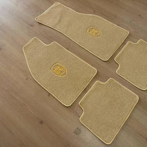 Puede incluir: Un juego de cuatro alfombrillas de coche de color beige con una superficie de moqueta texturizada. Cada alfombrilla presenta un emblema de escudo bordado en el centro en un tono beige a juego. El juego incluye dos alfombrillas delanteras más grandes y dos traseras más pequeñas.