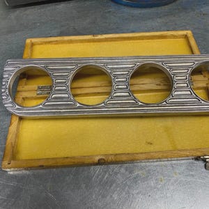 Pode incluir: Um painel de instrumentos retangular de metal prateado com quatro aberturas circulares dispostas horizontalmente. O painel apresenta um design texturizado e estriado entre cada abertura e está posicionado sobre uma caixa de madeira plana.