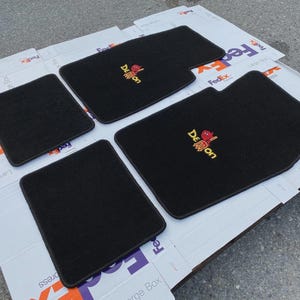 Puede incluir: Un juego de cuatro alfombrillas negras para coche con esquinas redondeadas. Dos alfombrillas presentan un logotipo bordado con un personaje de dibujos animados rojo y el texto amarillo "Demon". Están hechas de un material de moqueta oscuro y denso.